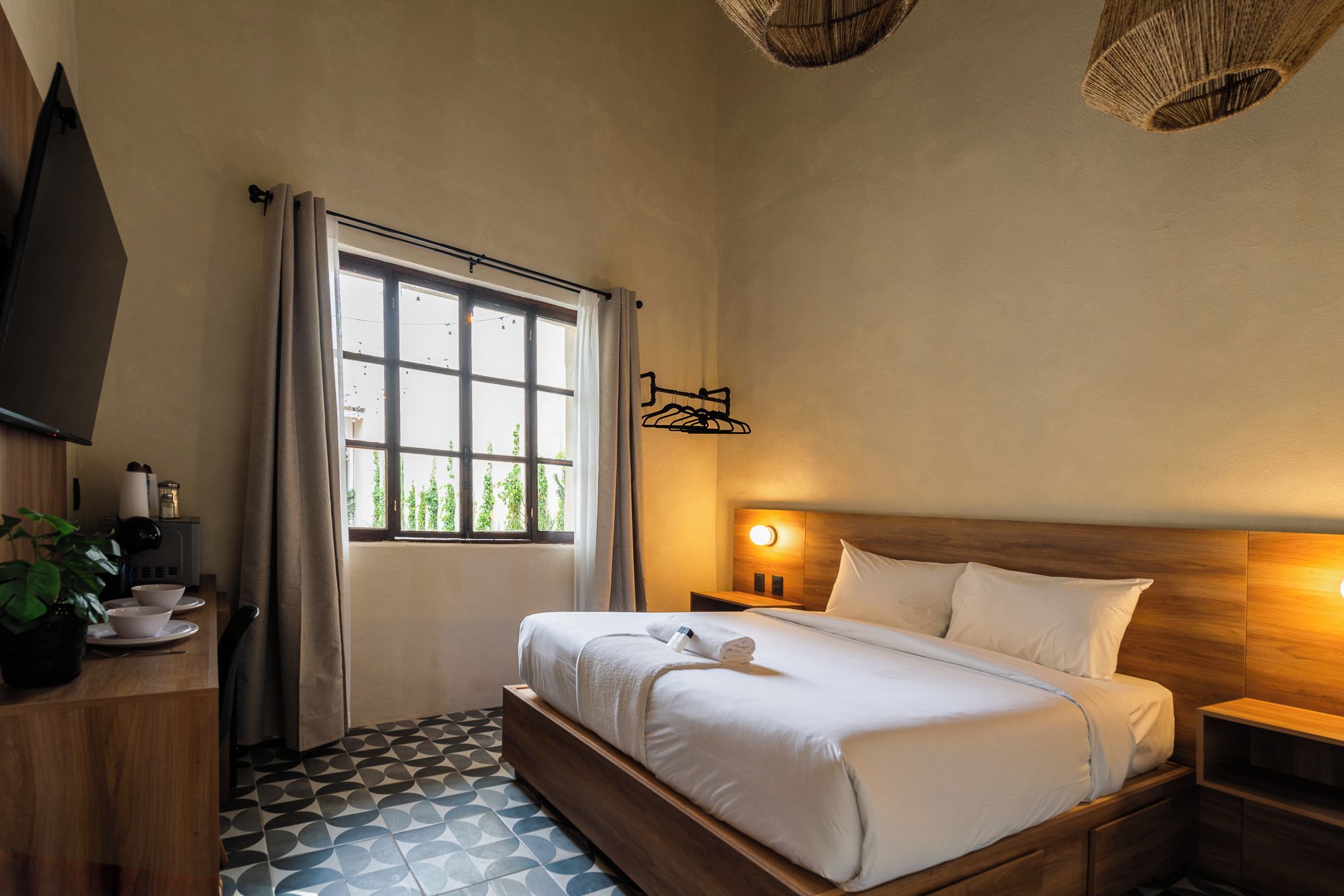 Habitación de hotel con cama, mesita de noche, televisor montado en la pared, ventana con cortinas y piso estampado.