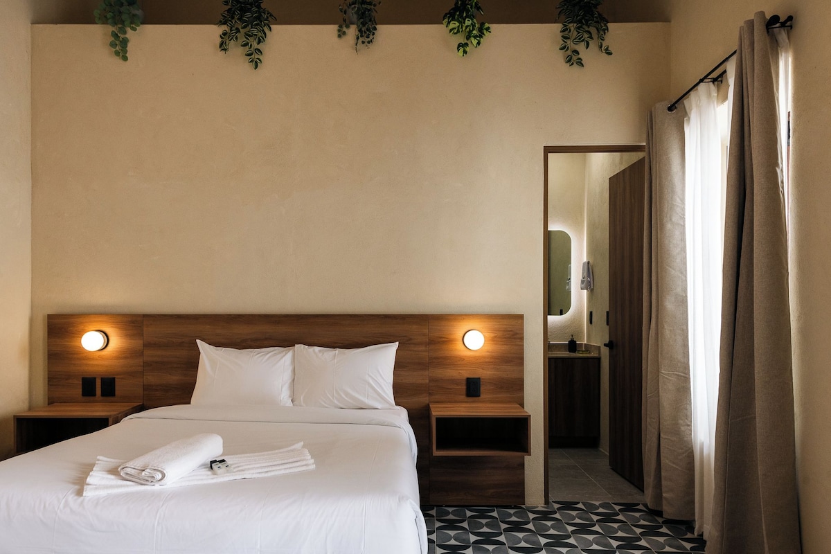 Una habitación de hotel con cama, mesitas de noche, un espejo y la puerta del baño.