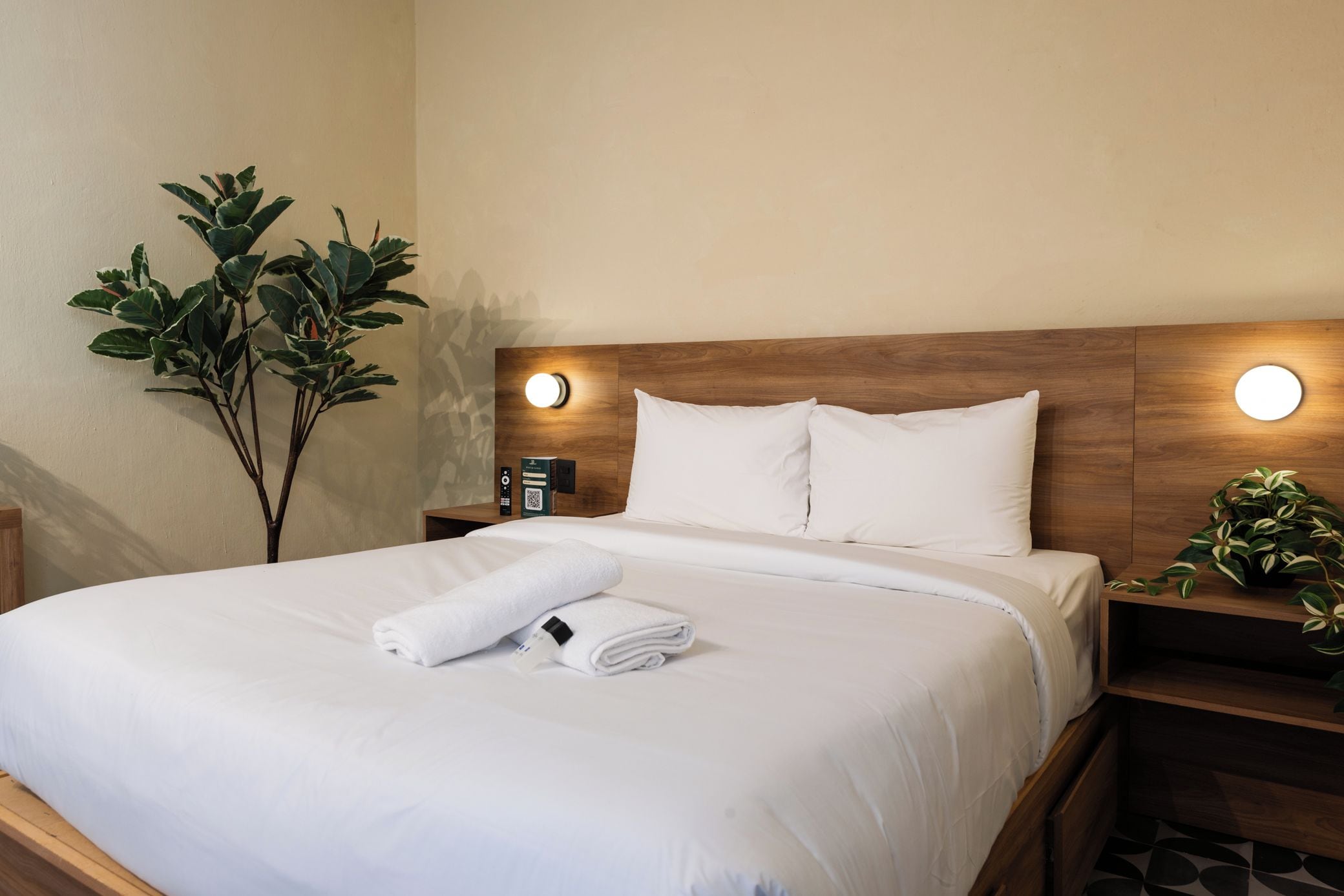 A neatly made bed with white linens, a folded towel, and a bedside table with a remote control and a book.