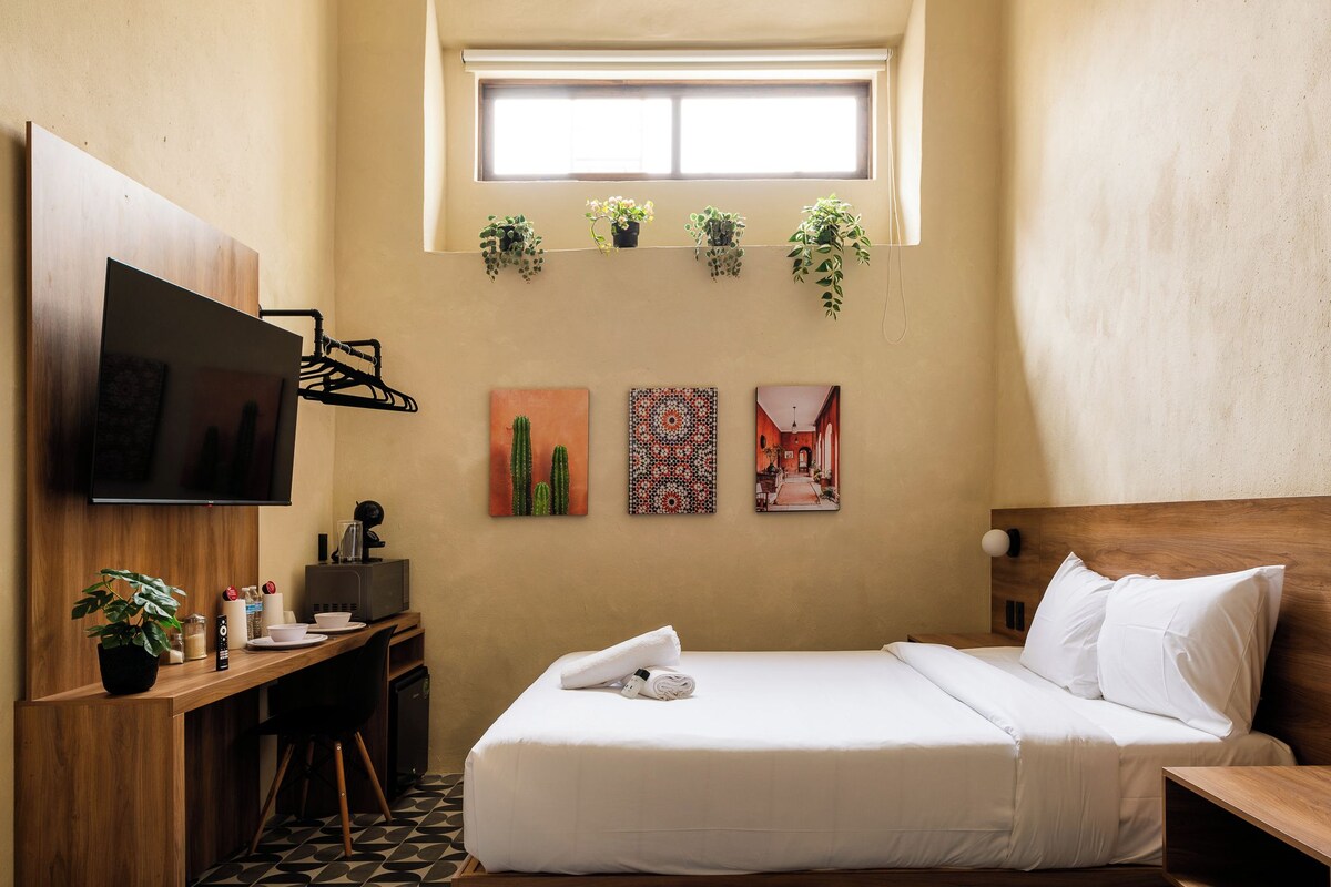 Una habitación de hotel con cama, escritorio con televisor, ventana con plantas y una obra de arte en la pared.