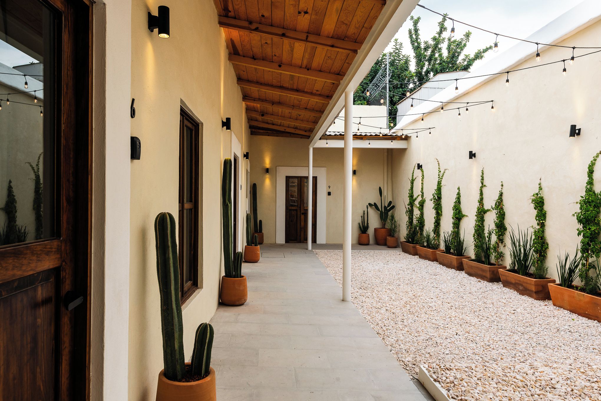 Un corredor cubierto con cactus y plantas en macetas, techo de madera y un edificio con el número '9' en la pared.