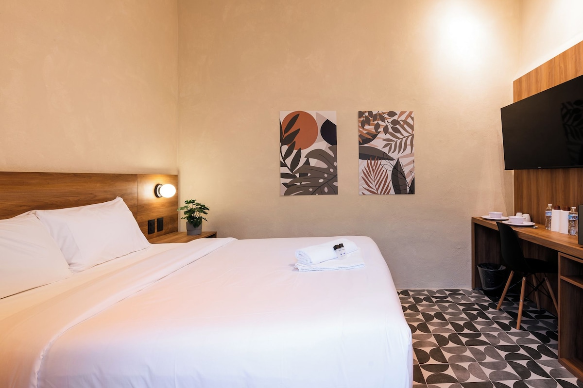 Habitación de hotel con una cama bien hecha, un escritorio con silla, una planta en maceta y dos obras de arte en la pared.