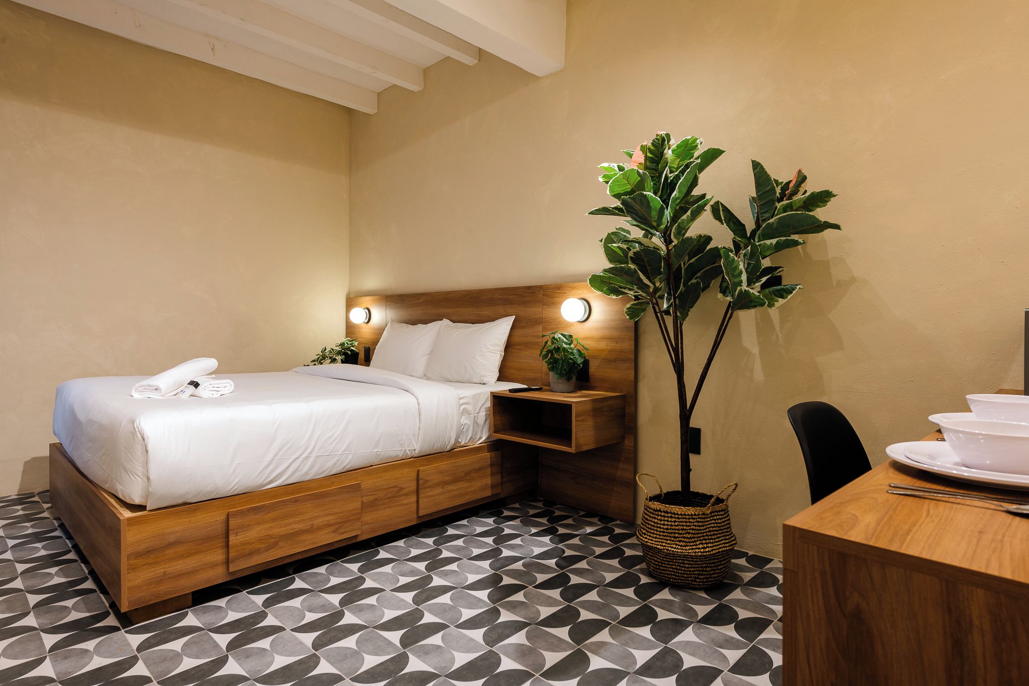 Habitación de hotel con cama de madera, una mesita de noche con lámpara, una planta en maceta y un comedor con mesa y sillas.