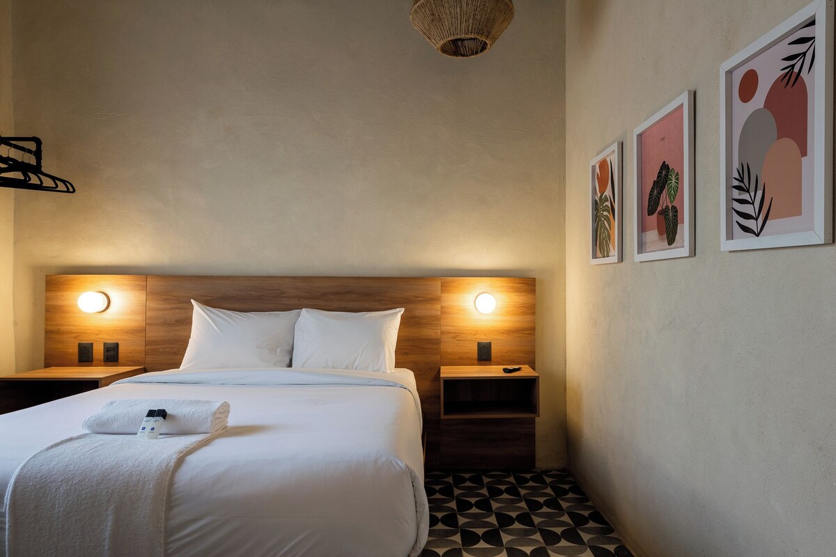 Habitación de hotel con cama, dos mesitas de noche, lámpara de pared e ilustraciones enmarcadas.