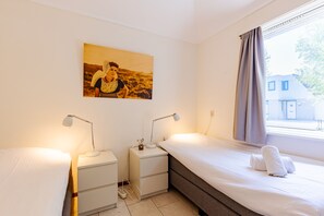 Ein Schlafzimmer mit zwei Einzelbetten, Nachttischen mit Lampen, einem Wandbild und einem Fenster mit Vorhängen.