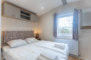 Un dormitorio con cama, mesita de noche con lámpara, ventana con cortinas y estanterías empotradas sobre la cama.