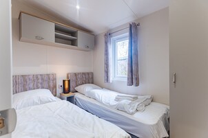 A bedroom with two single beds, a bedside table with a lamp, a window with curtains, and a built-in shelf above the beds.