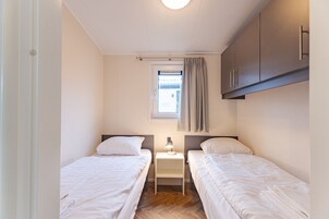 Un dormitorio pequeño y bien iluminado con dos camas individuales, una mesita de noche con lámpara, una ventana con cortinas y un armario.
