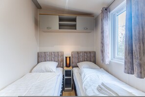 A small, compact bedroom with two single beds, a bedside table with a lamp, and a window with curtains. - Duinhoeve Chalet Schelp (Burgh-Haamstede)