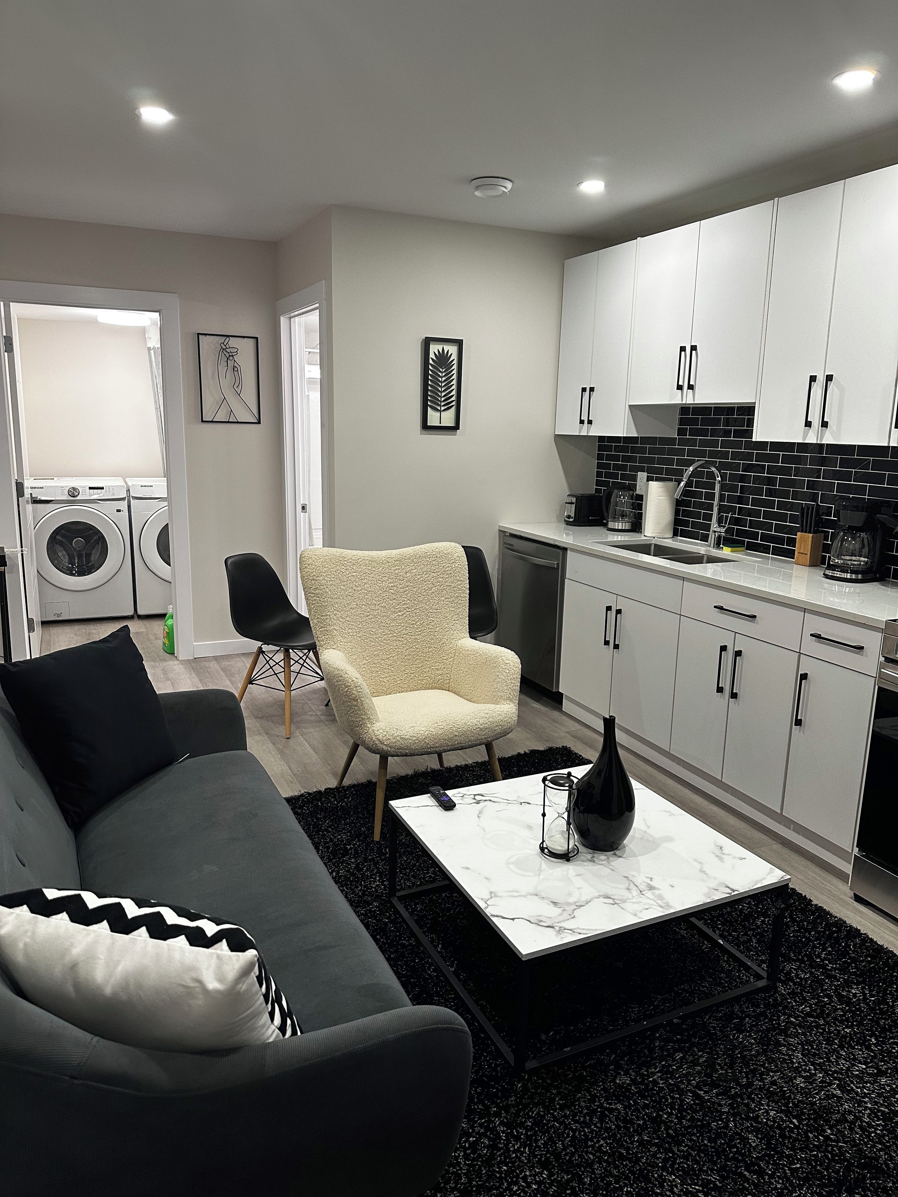 A modern living room with a sofa, armchair, and coffee table. A kitchen with white cabinets and black appliances is visible in the background.
