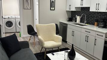 A modern living room with a sofa, armchair, and coffee table. A kitchen with white cabinets and black appliances is visible in the background.