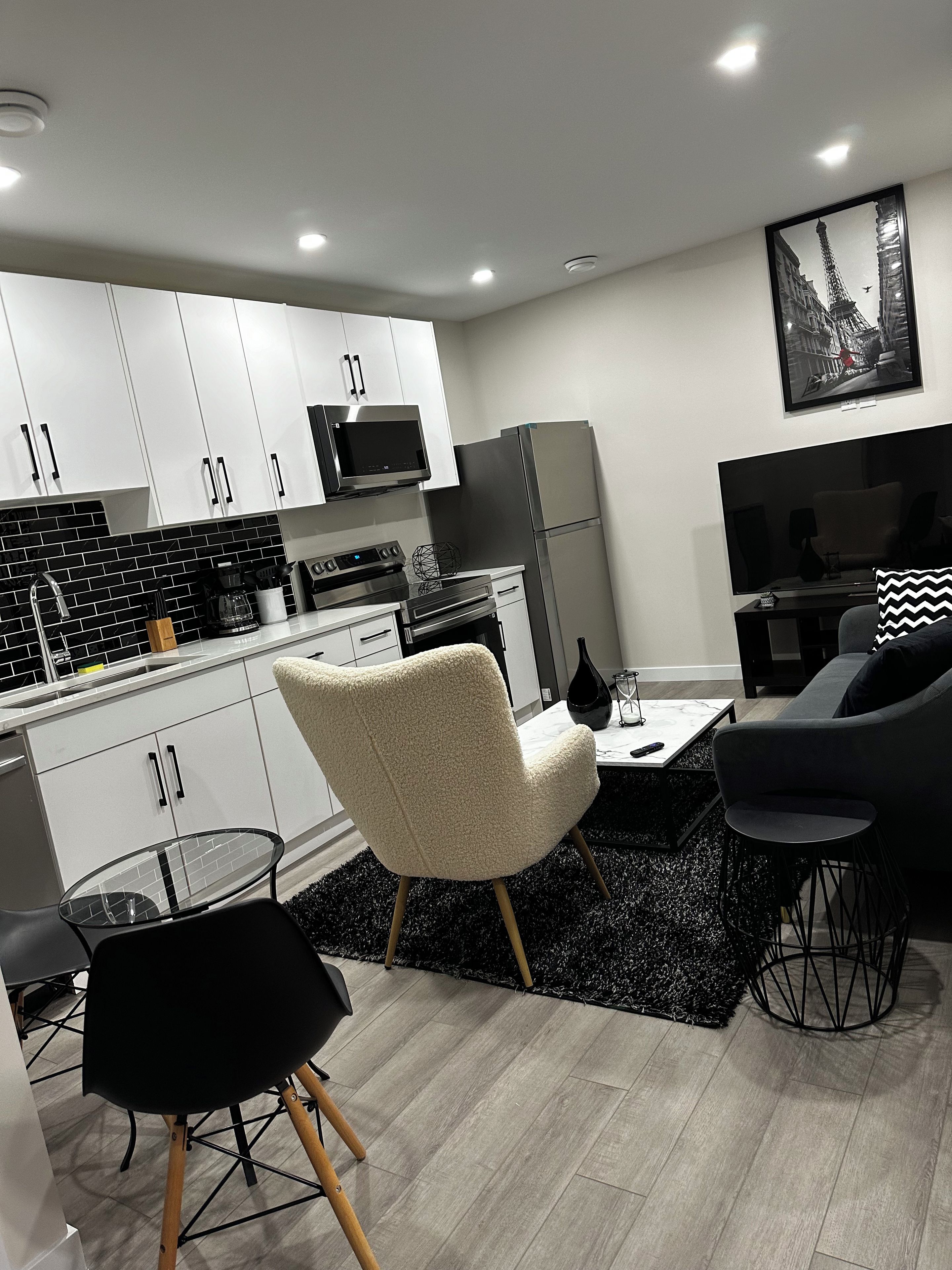 A modern kitchen with white cabinets, a microwave, and a refrigerator. There is a dining area with a black and white chair, a small table, and a black rug.