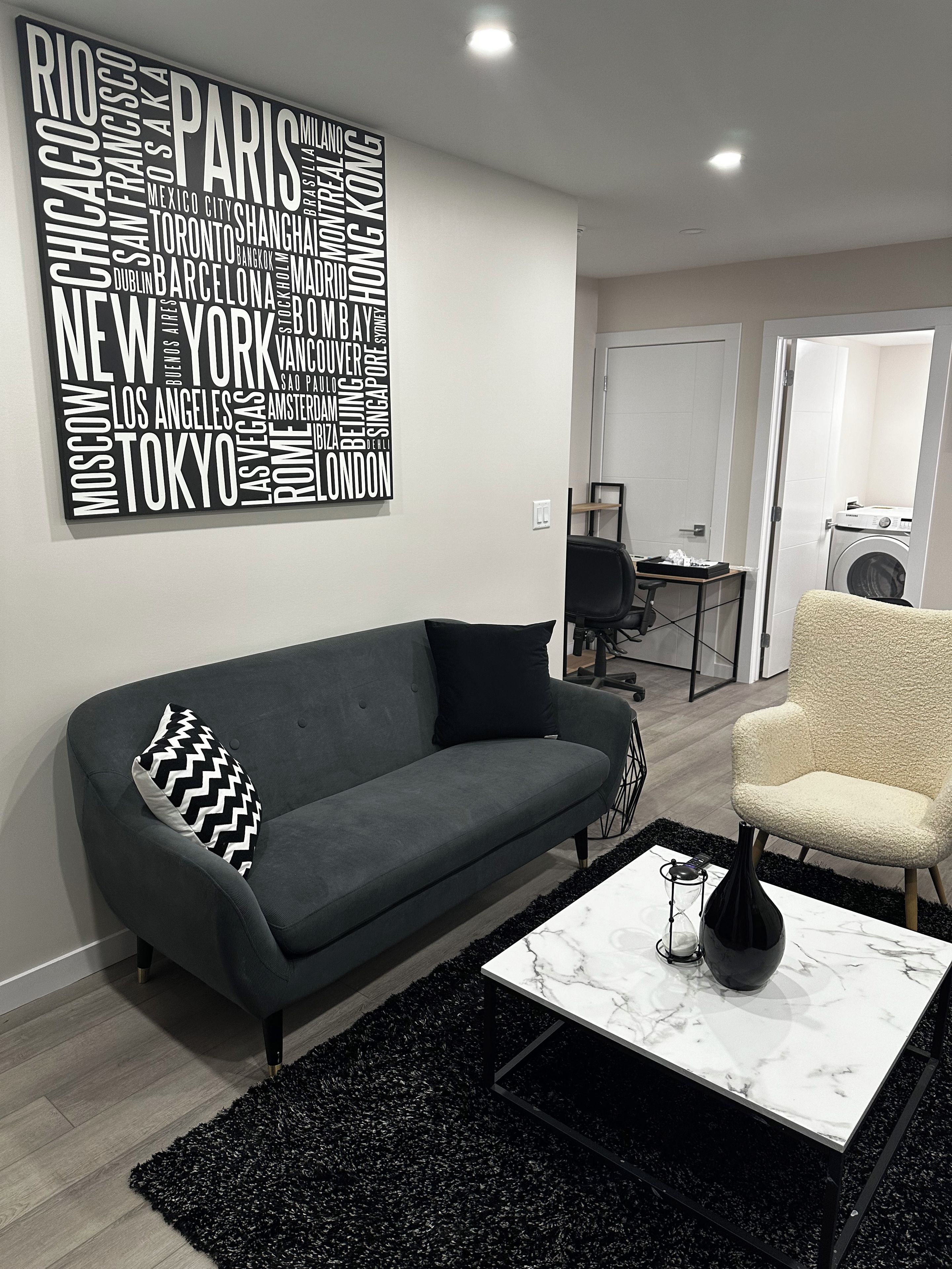 Uma sala de estar moderna com um sofá cinza, uma almofada estampada em preto e branco, uma mesa de centro de mármore branco e uma obra de arte com palavras na parede mostrando nomes de cidades.