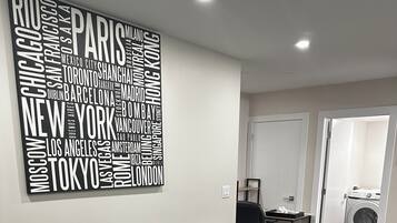 Uma sala de estar moderna com um sofá cinza, uma almofada estampada em preto e branco, uma mesa de centro de mármore branco e uma obra de arte com palavras na parede mostrando nomes de cidades.