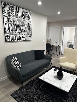 Un salon moderne avec un canapé gris, un coussin aux motifs noir et blanc, une table basse en marbre blanc et une décoration murale représentant un nuage de mots composé de noms de villes.