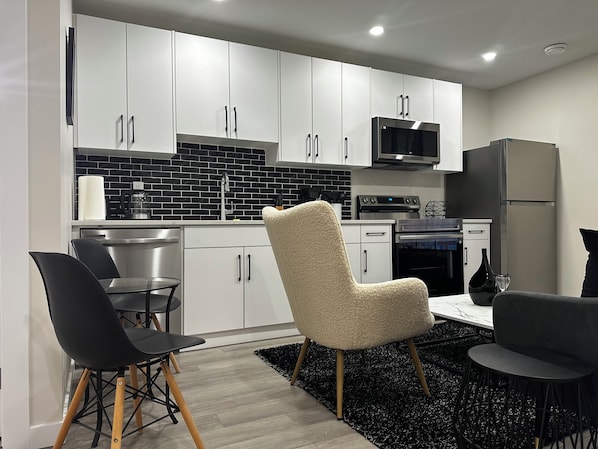 Une cuisine moderne avec des meubles blancs, un crédence en carreaux noirs, des appareils en acier inoxydable et un coin repas avec des chaises noires et un fauteuil beige.