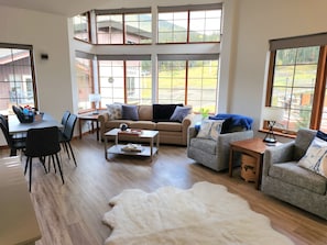 A spacious living room with a high ceiling, large windows, a sofa set, a coffee table, and a dining area with chairs.
