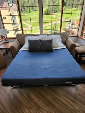 A bed with a blue cover, two pillows, and a view of a lodge and greenery through large windows.