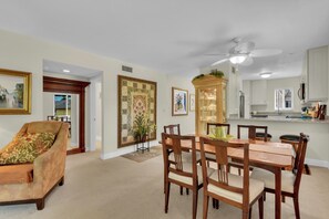 A dining area with a wooden table and chairs, a ceiling fan, and a kitchen with white cabinets and a window. - Olde Naples Getaway | 2BR Condo Near Beach & 3rd Ave S (Naples)