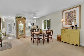 Een eetgedeelte met een houten tafel en stoelen, een plafondventilator, een keuken met witte kasten en een geel kastje met een lamp.
