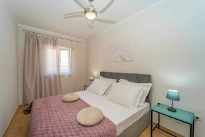 A bedroom with a bed, a ceiling fan, a window with curtains, a bedside table with a lamp, and a wall-mounted artwork.