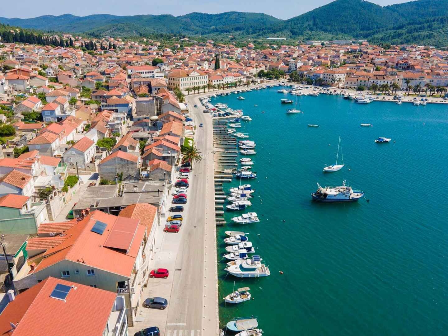 A coastal town with a marina filled with boats, a beach, and buildings with red-tiled roofs.