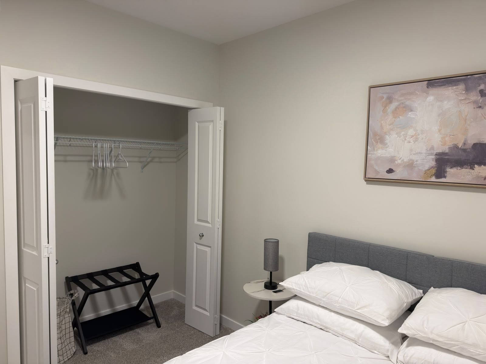 A bedroom with a bed, a bedside table with a lamp, a closet with hanging clothes, and a wall-mounted abstract painting.