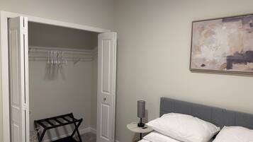 A bedroom with a bed, a bedside table with a lamp, a closet with hanging clothes, and a wall-mounted abstract painting.