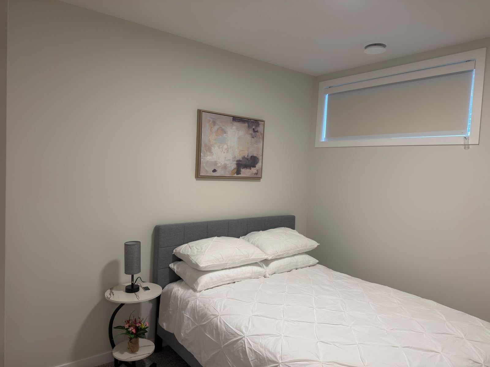 A bedroom with a bed, a bedside table with a lamp, a small plant, and a painting on the wall.