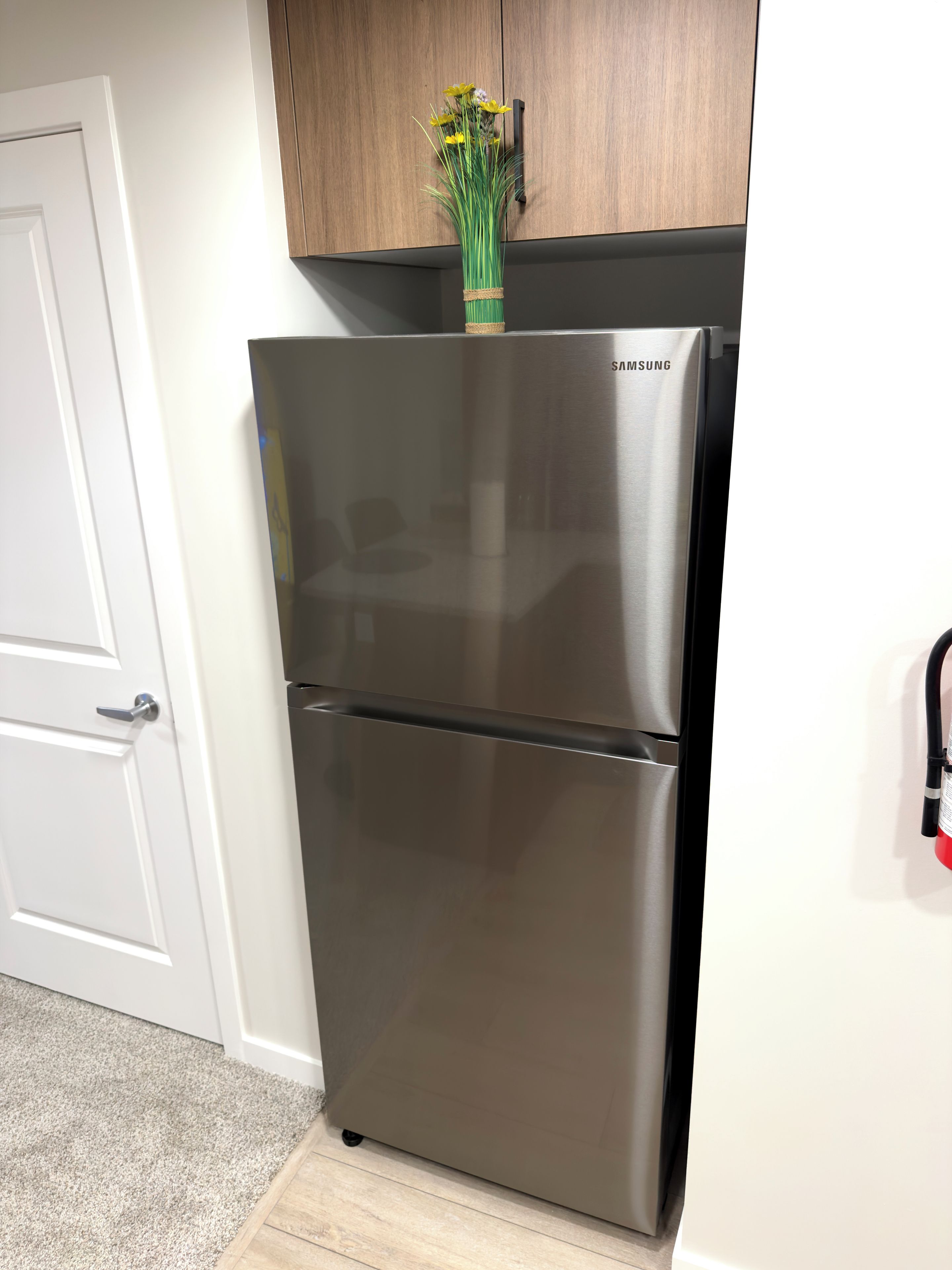 A stainless steel refrigerator with a Samsung logo, a wooden cabinet above it, and a white door to the left.