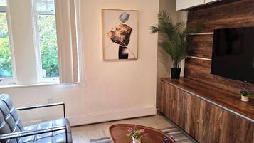 A modern living room with a leather sofa, a wooden coffee table with a remote, a framed abstract painting, a large TV, and a potted plant.