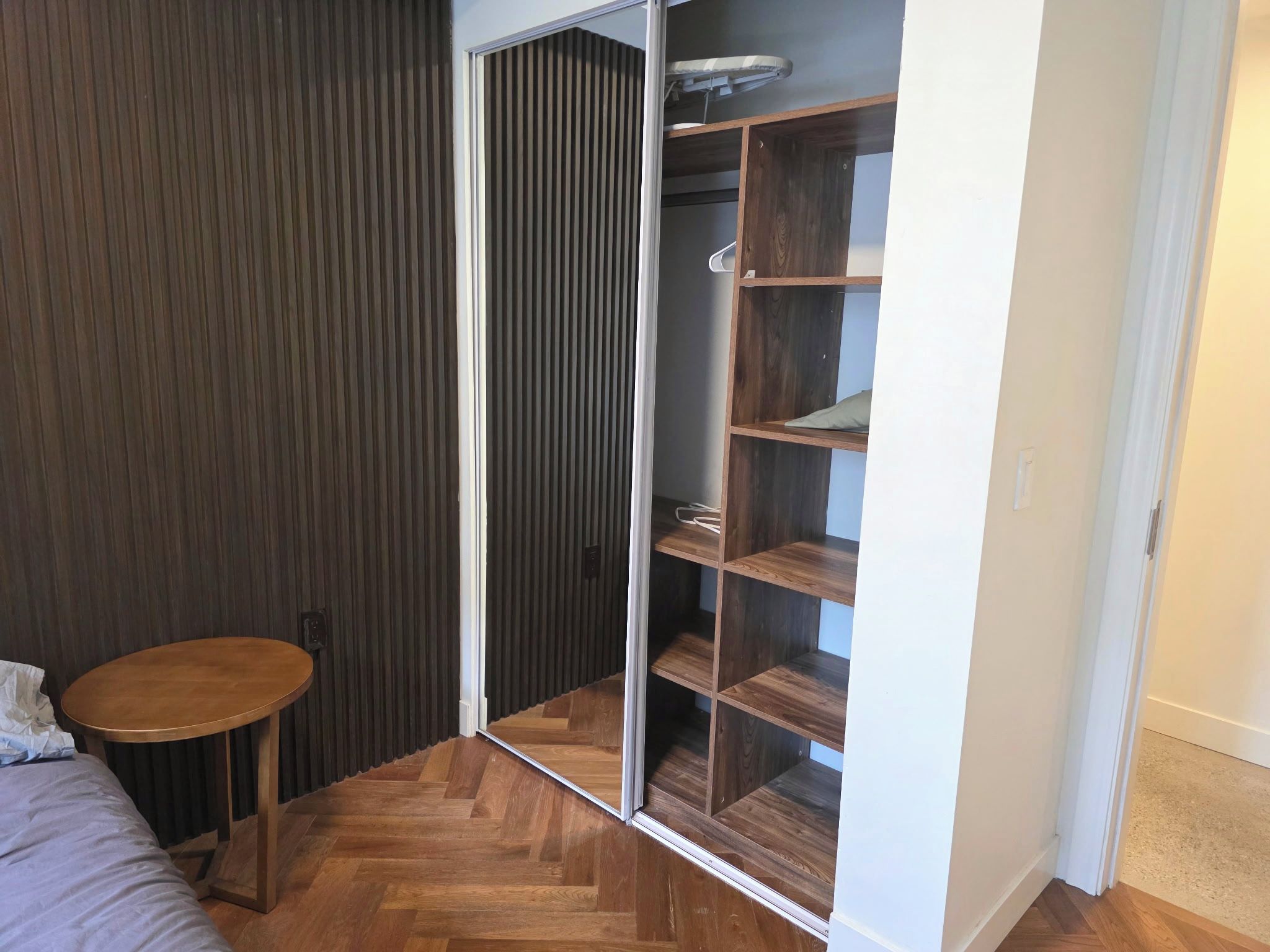 A room with a wooden herringbone floor, a small round table, a wardrobe with multiple shelves, and a sliding door.