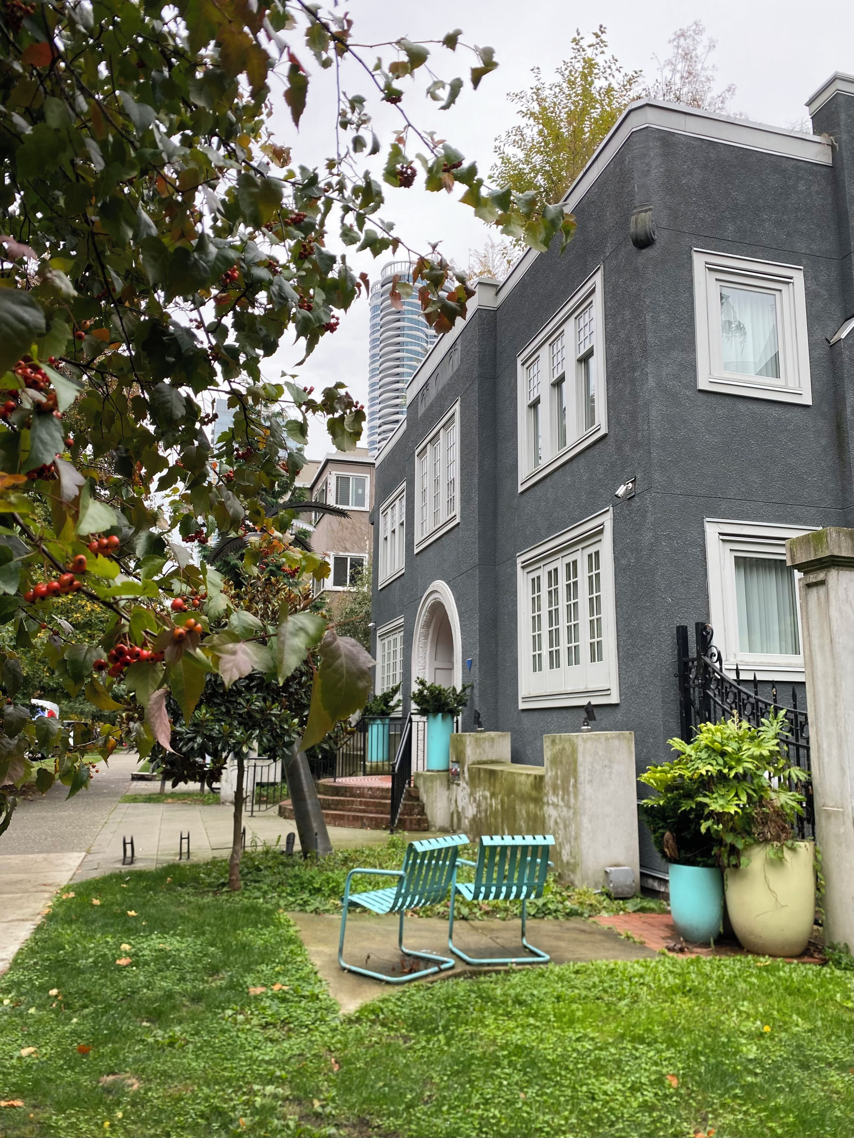 A building with a grey facade, white windows, and a small garden with a bench and potted plants.