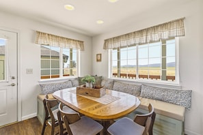 A dining area with a round wooden table, cushioned seating, and a view of a landscape through large windows.