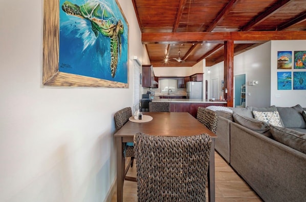 A dining area with a wooden table and woven chairs, a kitchen in the background, and a large painting of a sea turtle on the wall. - Cozy 2-bedroom Zen style house in peaceful Roatan Bay Island (Roatan Bay Island)
