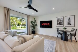 A modern living room with a sofa, a dining table with chairs, a ceiling fan, a large window with curtains, a potted plant, and a wall-mounted TV displaying 'NETFLIX'. - Chic 2BR on South Congress / Private Backyard (Austin)