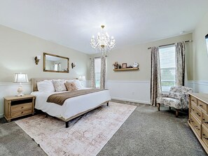 Ein Schlafzimmer mit Bett, Nachttisch, Kommode, einer Kronleuchter und einem Fenster mit Vorhängen.