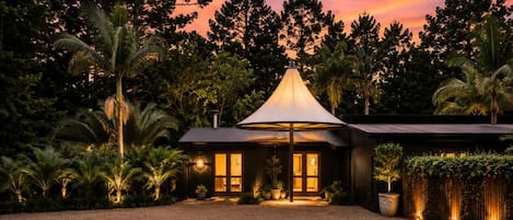 A modern house with a large umbrella-like structure in front, surrounded by lush greenery and palm trees.
