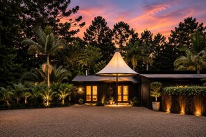 Una casa moderna con una gran estructura en forma de paraguas al frente, rodeada de exuberante vegetación y palmeras.