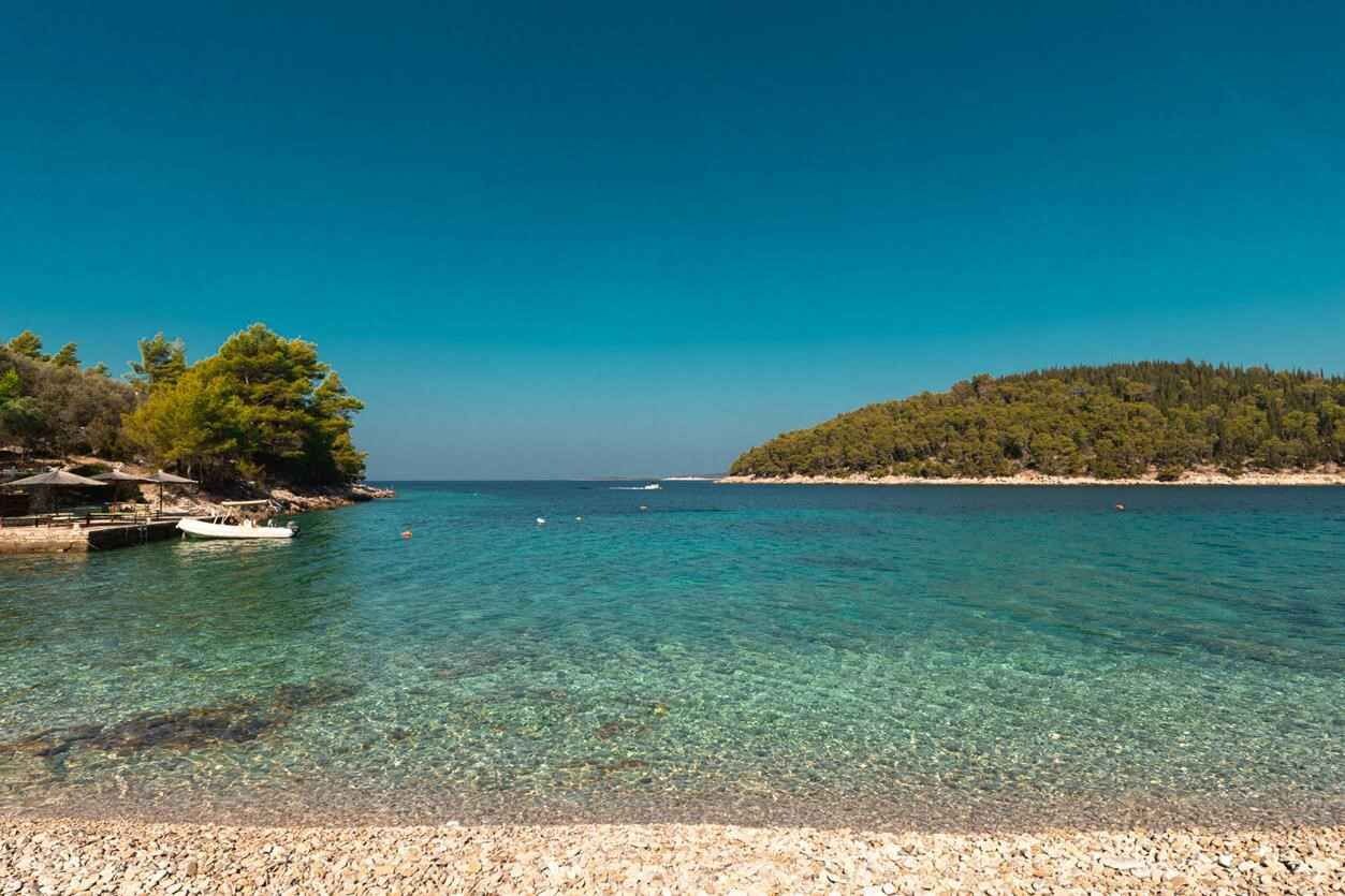 A clear coastal view with a pebble beach, a boat, and a forested island.