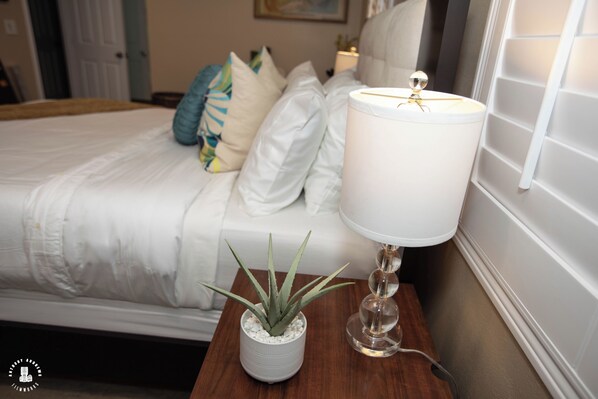Una cama bien hecha con ropa de cama blanca, una lámpara de noche y una pequeña planta en maceta sobre una mesita de madera.