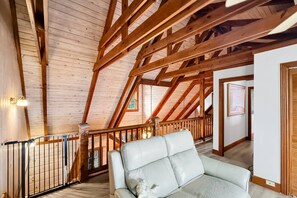 A high-ceilinged room with exposed wooden beams, a cushioned seating area, and a staircase leading to another level. - Free Ruby Falls Ticket | Mountain View Log Cabin | Mini Golf + Fire Pit | 5 minutes Cloudland Canyon (Rising Fawn)