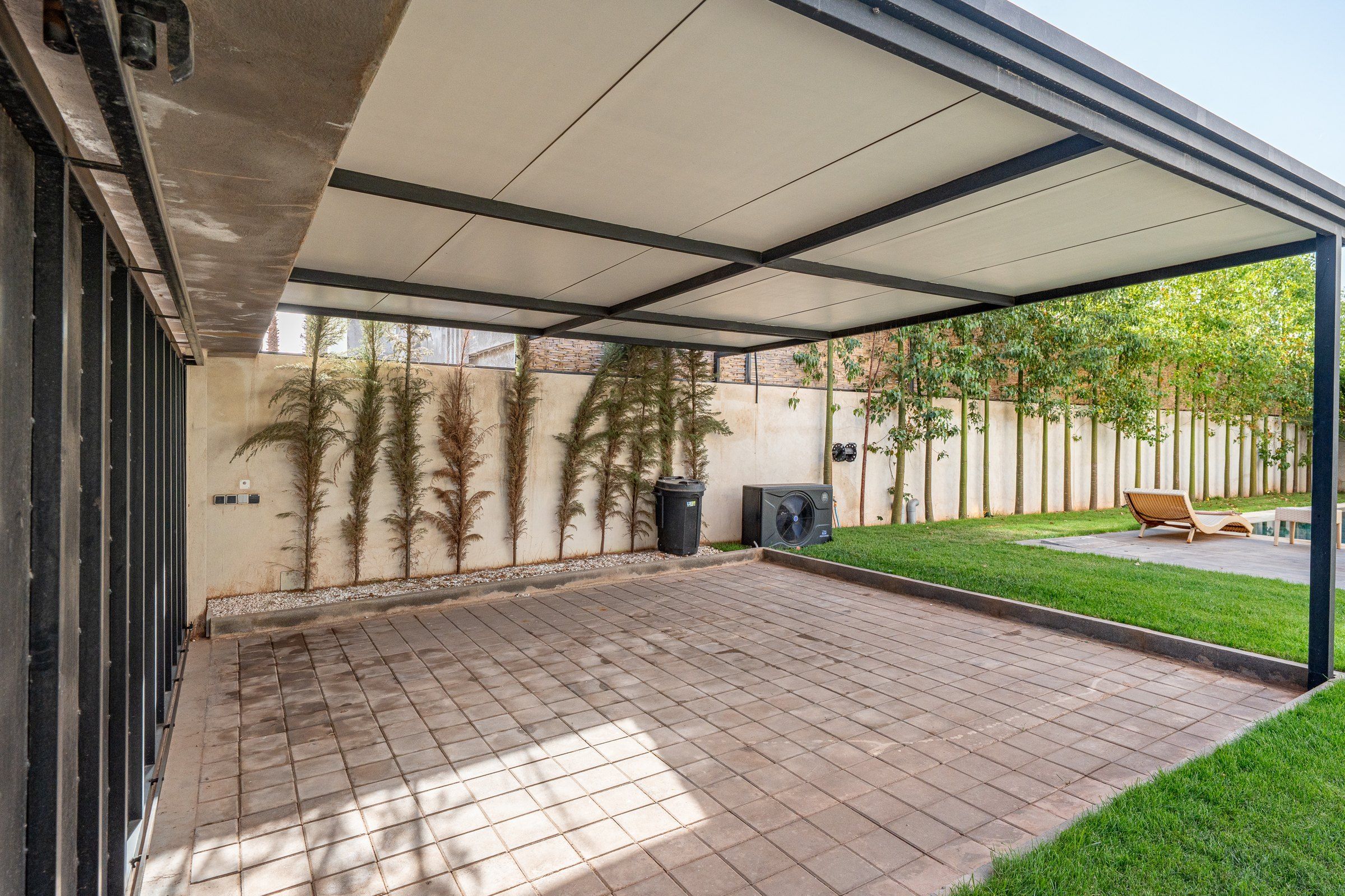 Ein überdachter Patio-Bereich mit Fliesenboden, einer mit Kletterpflanzen bewachsenen Wand und einem Rasen mit Pool und Liegestühlen.