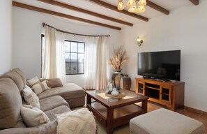 A living room with a sofa, a coffee table, a TV stand, and a window with curtains. - Villa Europa: Private Pool and Space for Families and Groups (Nazaret)