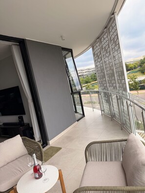 Un balcó modern amb barana de vidre, un televisor de pantalla plana i una zona d'estar amb taula i cadires.