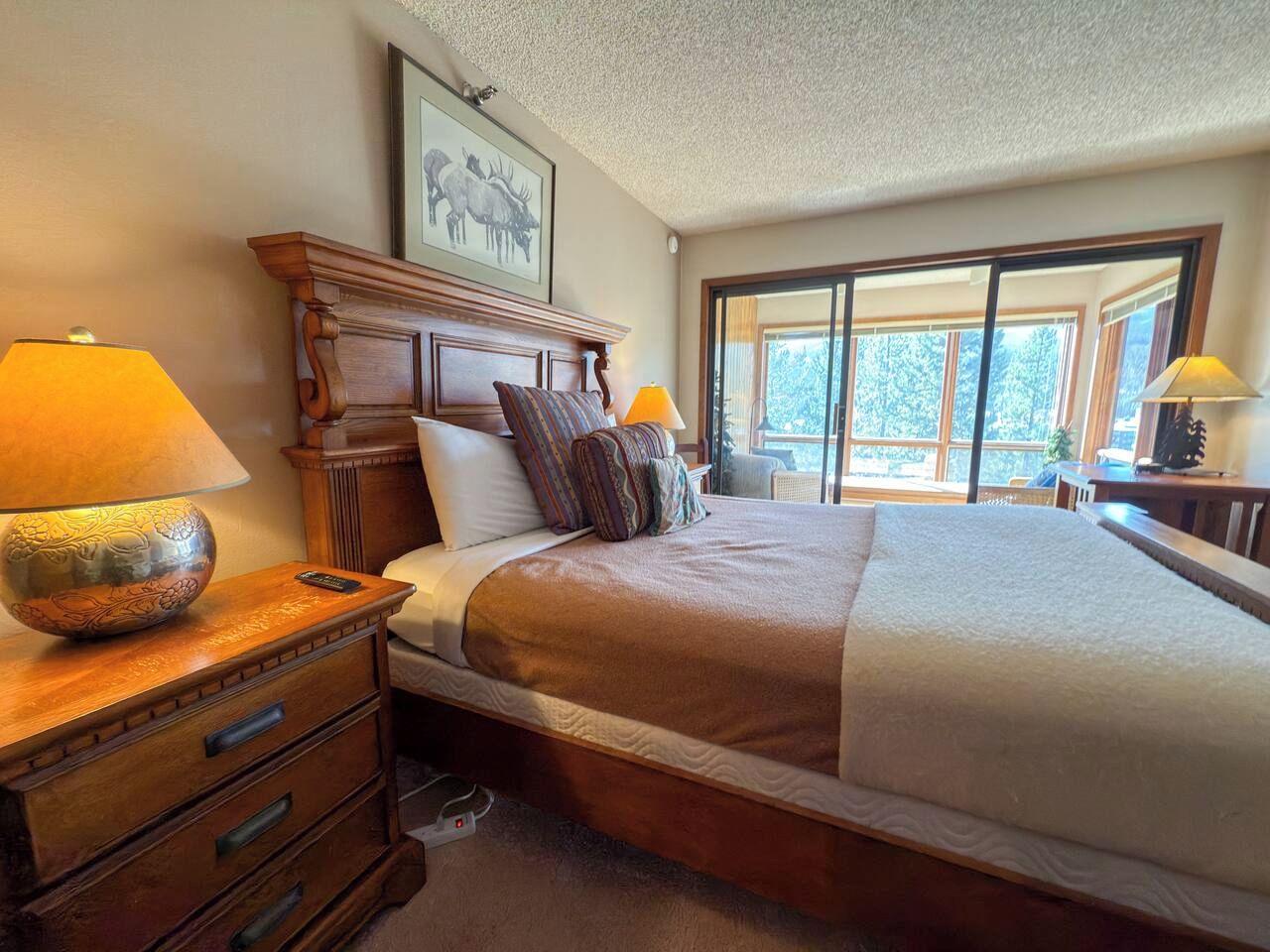 A hotel room with a large bed, wooden headboard, two bedside lamps, a nightstand, and a window with a view of trees.