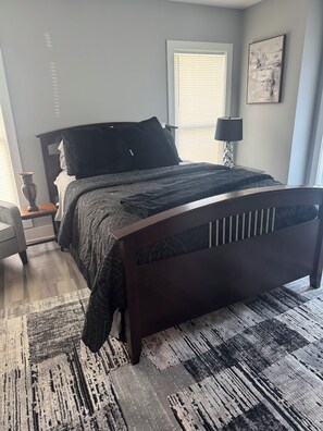 A bedroom with a bed, a nightstand, a lamp, a window with blinds, and a painting on the wall. - Just Like New-Modern 3 Bedroom House Near HOTLANTA (Decatur)