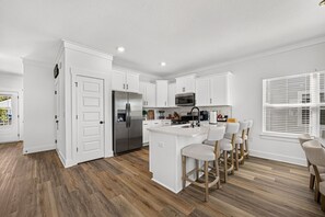A modern kitchen with white cabinetry, a stainless steel refrigerator, a microwave, and a bar area with stools. - New home—5 minutes from the beach, Pier Park, and Walmart; includes an arcade room, balcony, and work area*pets allowed (Panama City beach)