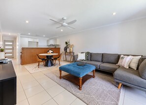 A modern living room with a grey sofa, a blue ottoman, a dining table with chairs, and a kitchen area in the background. - Caloundra Townhouse  2 bed, 2 bath, 2 car, 5 mins to beaches. Private (Caloundra West)