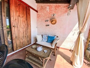 A small patio area with a wicker table, a sofa with patterned cushions, a wooden door, and a wall with a textured finish. - Nice apartment for 6 people with A/C, pool and patio (Olbia)